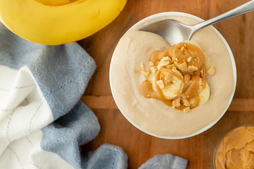 A bowl of creamy banana smoothie topped with banana slices, chopped nuts, and peanut butter, perfect for high protein snacks. A banana and a blue and white cloth napkin are nearby on a wooden surface, with a spoon ready to enjoy.