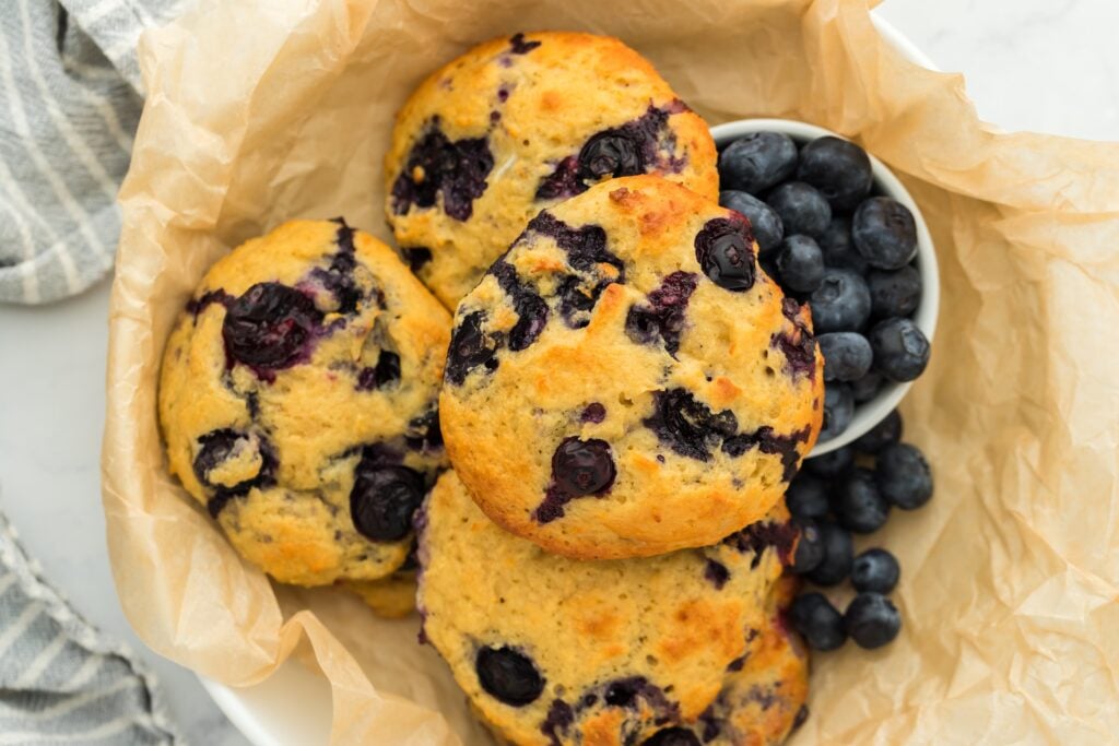 Three golden-brown blueberry muffins, perfect as high protein snacks, rest in a parchment-lined bowl beside a small white cup filled with fresh blueberries. A striped cloth is partially visible at the edge.