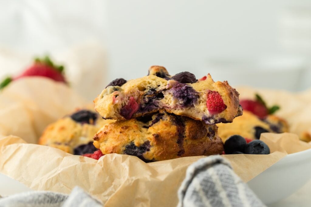 A close-up of freshly baked, high protein berry scones, with visible strawberries and blueberries, resting on parchment paper. One scone is broken open, showing its moist, berry-filled interior. A gray cloth is in the foreground.