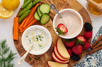 A wooden platter with sliced apples, strawberries, blackberries, carrot sticks, snap peas, cucumber, and two bowls of dip—one creamy white and one light brown—garnished with herbs and berries. Perfect for those seeking high protein snacks. A lemon and honey dipper are nearby.