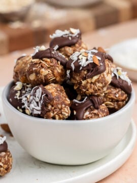 A white bowl filled with chocolate-dipped oat and nut energy bites topped with shredded coconut, surrounded by whole almonds and a striped towel on a light surface.
