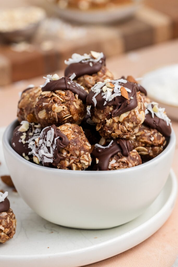 A white bowl filled with chocolate-dipped oat and nut energy bites topped with shredded coconut, surrounded by whole almonds and a striped towel on a light surface.