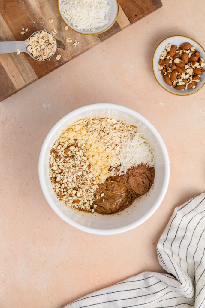 A white bowl contains oats, shredded coconut, chopped nuts, cocoa powder, and nut butter. Nearby are bowls with more nuts and coconut, a measuring cup with oats, a cutting board, and a striped kitchen towel.