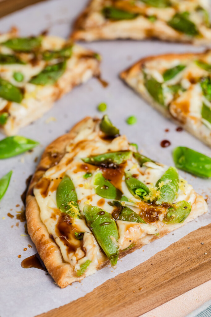 A close-up of a slice of pizza topped with snap peas, melted cheese, and a drizzle of dark sauce, resting on parchment paper with other slices nearby.