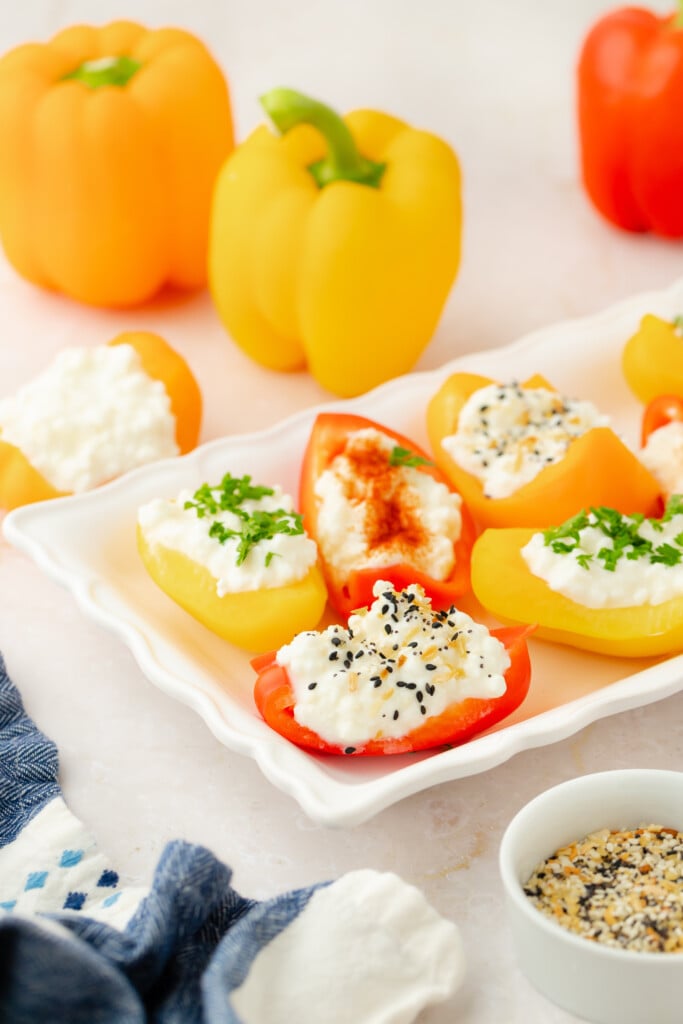 A white plate holds sliced yellow and red bell peppers filled with cottage cheese, topped with chives, paprika, and everything bagel seasoning. Whole peppers and a bowl of seasoning are in the background.