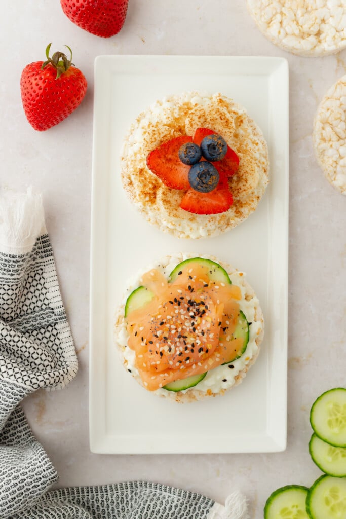 A rectangular white plate with two rice cakes: one topped with sliced strawberries and blueberries, the other with cucumber slices, smoked salmon, sesame seeds, and cream cheese. Strawberries, rice cakes, and cucumber slices are nearby.