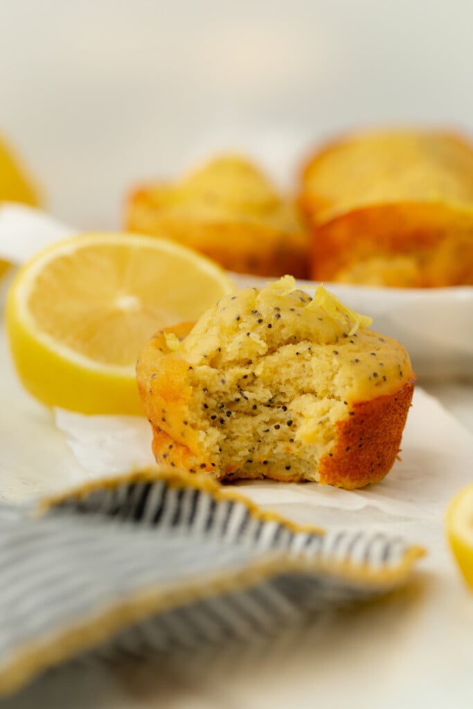 A Bakery-Style Lemon Poppy Seed Muffin with a bite taken out, surrounded by lemon halves and other muffins, sits on a white surface with a striped napkin in the foreground.