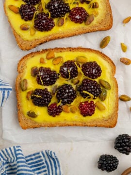 Two slices of Blackberry-Pistachio Custard Toast topped with lemon curd, blackberries, pistachios, and lemon zest sit on parchment paper, surrounded by a striped blue cloth, loose blackberries, pistachios, and a zested lemon.