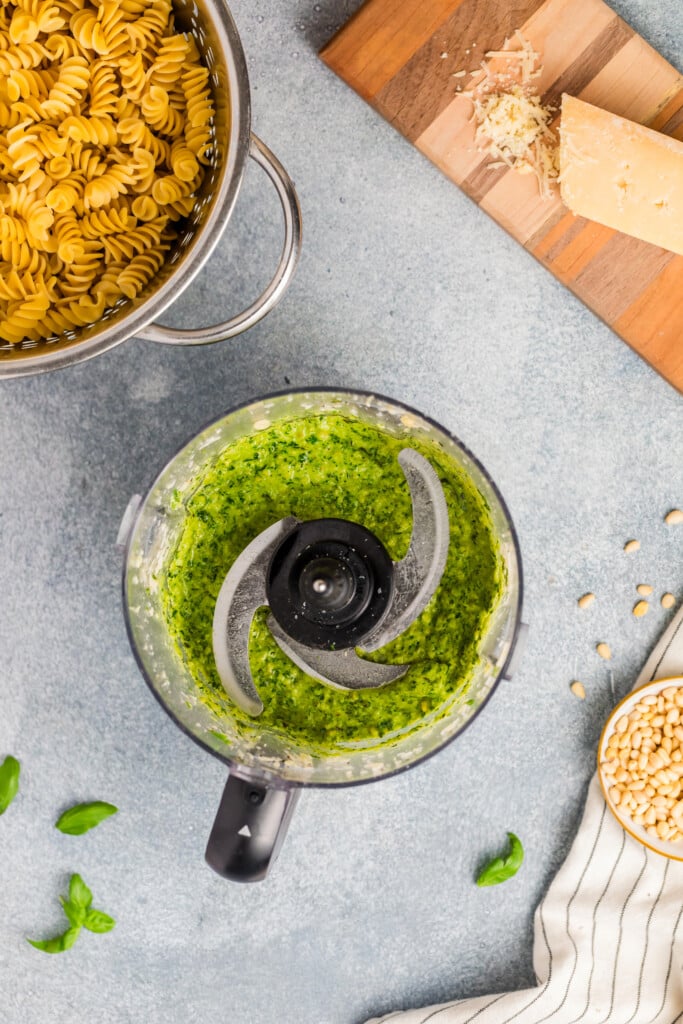 A food processor with green pesto sits on a countertop next to a pot of uncooked rotini, Parmesan cheese, pine nuts, and a striped towel—all the essentials for preparing a delicious Pesto Caprese Pasta Salad.