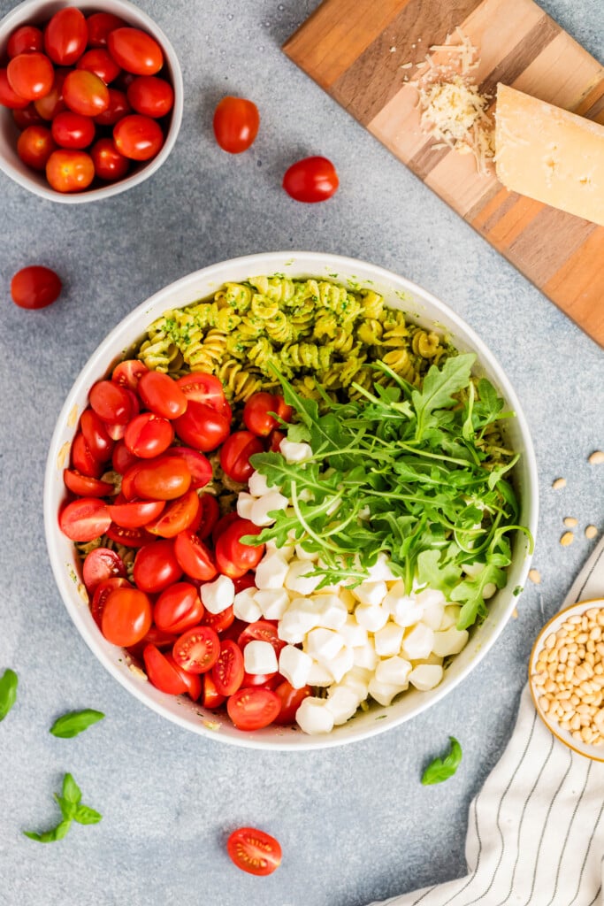 A bowl of Pesto Caprese Pasta Salad filled with pesto pasta, halved cherry tomatoes, fresh arugula, and mini mozzarella balls sits on a countertop, surrounded by cherry tomatoes, a cheese wedge, and scattered pine nuts and basil.