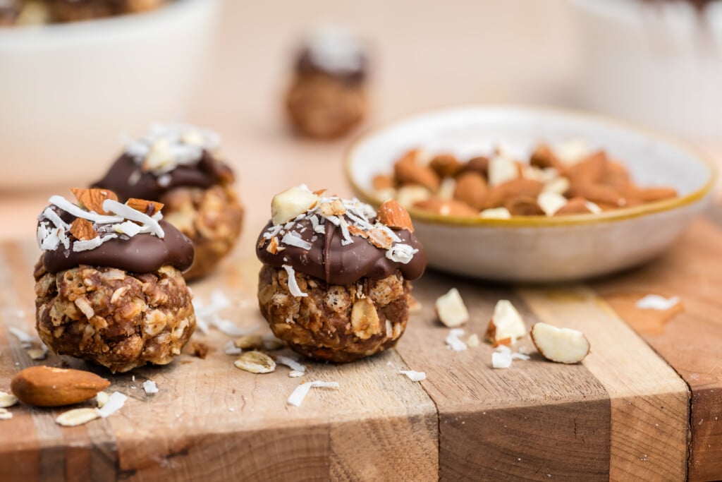 Three homemade energy bites topped with dark chocolate, chopped nuts, and shredded coconut sit on a wooden board, with a bowl of whole almonds and more bites blurred in the background.