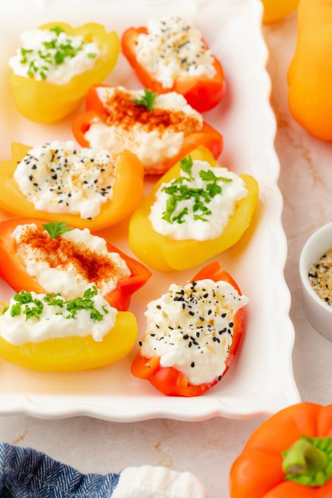 A white tray holds halved yellow and red mini bell peppers filled with cottage cheese and topped with various herbs, seeds, and spices. A bowl of seasoning and whole peppers are nearby on the table.