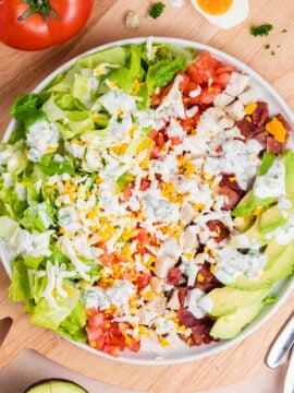 A bowl of Cobb salad with chopped lettuce, tomato, grilled chicken, bacon, avocado, shredded cheese, and creamy dressing, on a wooden surface with a halved boiled egg, tomato, and avocado nearby.