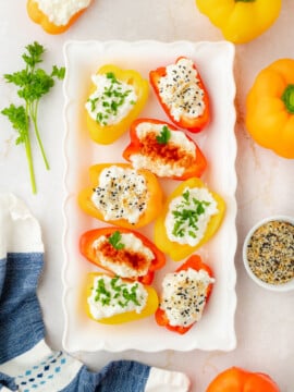 A white platter holds mini bell pepper halves filled with cottage cheese, topped with herbs and seasonings. Yellow and orange whole peppers, parsley, a spice bowl, and a blue cloth are nearby on a light surface.