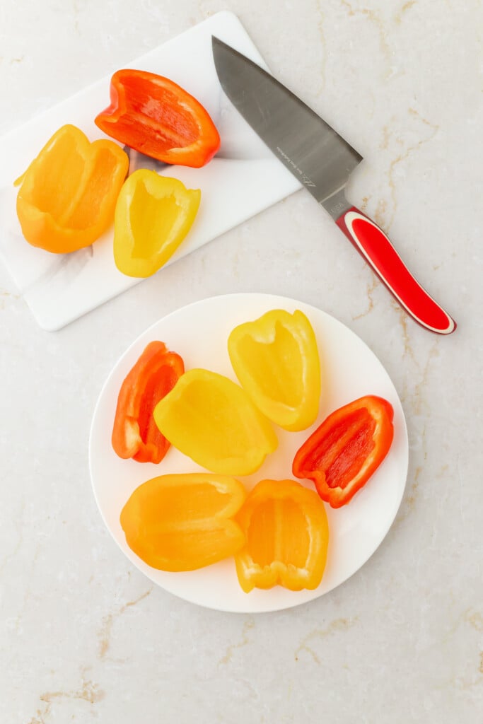 Sliced yellow and red bell peppers on a white plate, with more pepper halves and a knife with a red handle on a white cutting board, all on a light-colored countertop.