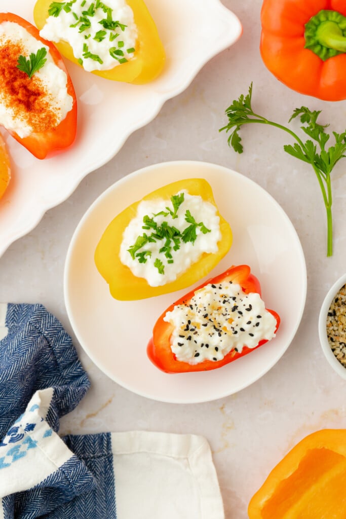 Halved yellow and red bell peppers filled with cottage cheese, topped with herbs and spices, arranged on a plate and tray, with a blue napkin and ingredients nearby on a light surface.