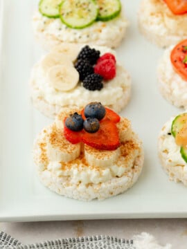 A row of rice cakes topped with cottage cheese and assorted fresh fruits, including blueberries, bananas, strawberries, blackberries, raspberries, and cucumber slices, arranged on a white rectangular plate.