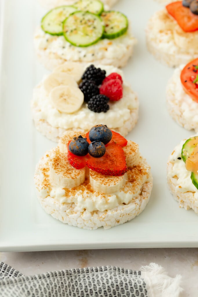 A row of rice cakes topped with cottage cheese and assorted fresh fruits, including blueberries, bananas, strawberries, blackberries, raspberries, and cucumber slices, arranged on a white rectangular plate.