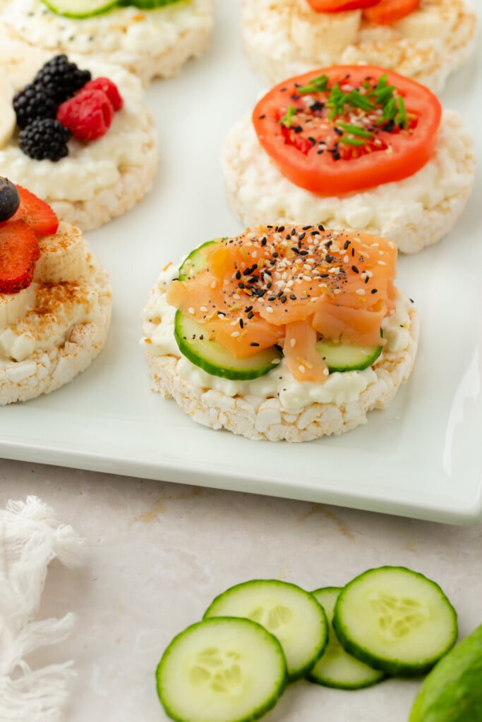 A white plate holds assorted rice cakes topped with various ingredients, including berries, tomato slices with herbs, and cucumber slices with smoked salmon and seasoning. Sliced cucumbers are arranged on the table nearby.