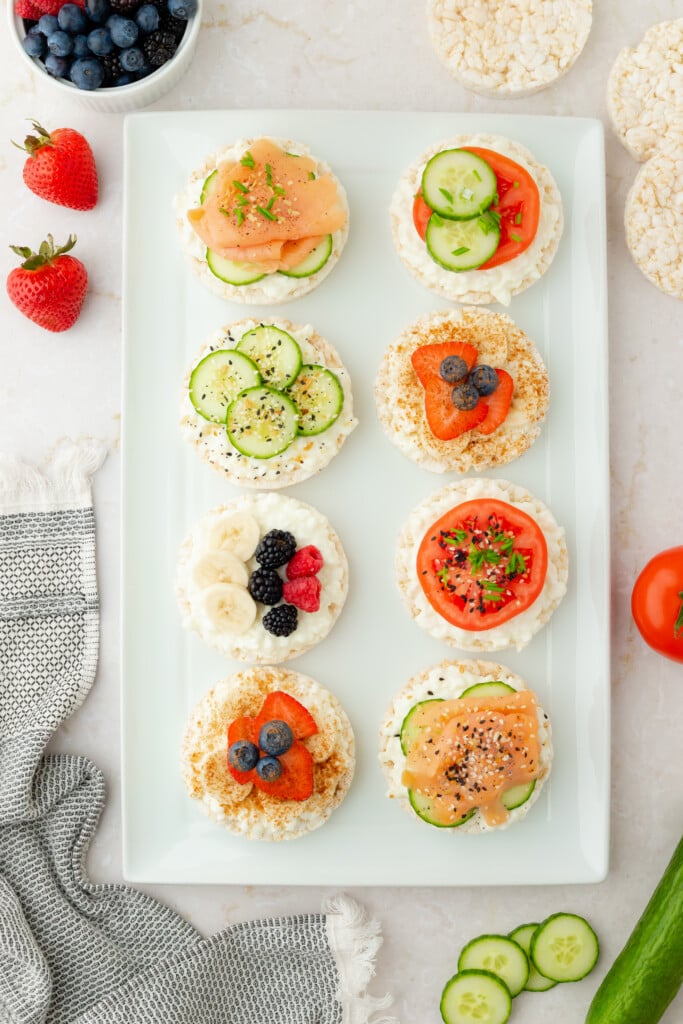 A white rectangular plate with eight rice cakes topped with various ingredients like smoked salmon, sliced cucumbers, tomatoes, berries, bananas, and cream cheese. Fresh fruit and veggies surround the plate on a marble surface.
