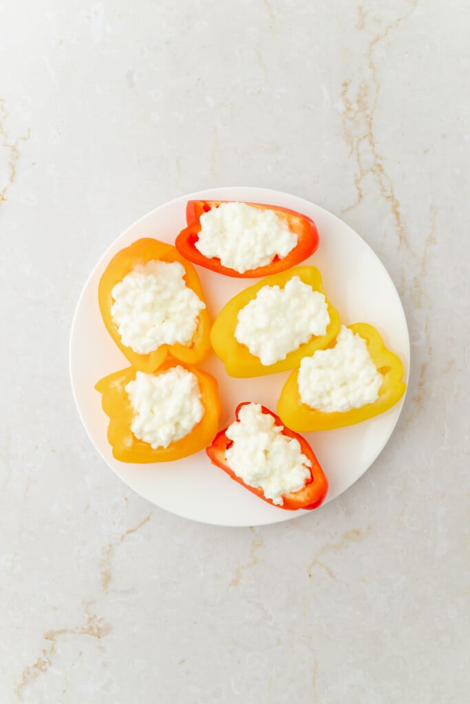 A white plate with six bell pepper halves, red and yellow, filled with cottage cheese, placed on a light marble surface.