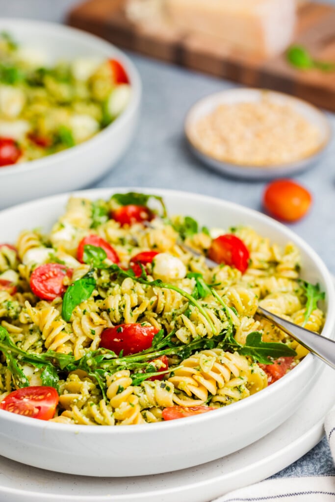 A white bowl filled with Pesto Caprese Pasta Salad—rotini, cherry tomatoes, mozzarella balls, arugula, and pesto. Another salad bowl and ingredients are blurred in the background. A fork rests on the side of the bowl.