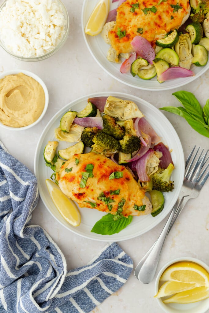 A plate of Roasted Garlic Hummus Chicken & Veggies (Easy One Sheet Dinner) featuring baked chicken breast, roasted broccoli, zucchini, red onion, and artichoke, garnished with basil and lemon. Served with hummus, feta, and a blue-striped napkin.