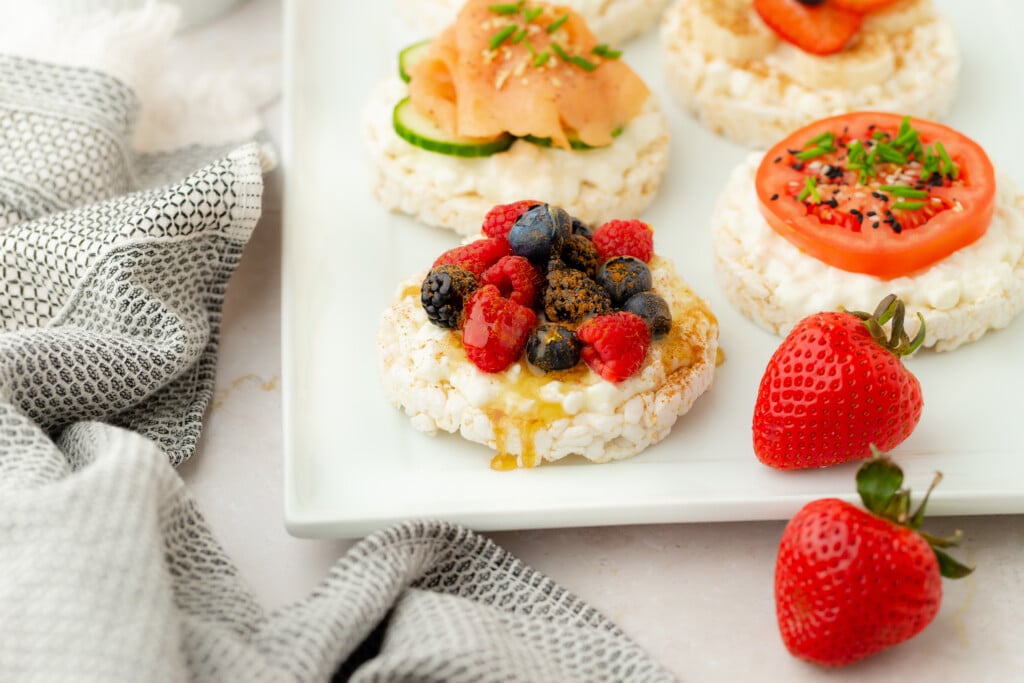 A plate with rice cakes topped with various ingredients: one with berries and honey, one with smoked salmon and cucumber, and one with tomato slices. Two fresh strawberries are on the side, next to a gray towel.