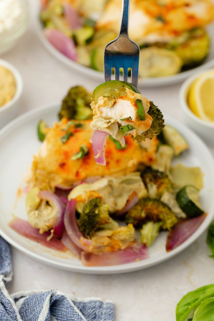 A fork holds a bite of Roasted Garlic Hummus Chicken & Veggies (Easy One Sheet Dinner) with baked chicken, broccoli, zucchini, and red onion over a plate of more roasted vegetables and chicken. Lemon wedges and a blue cloth napkin are nearby.