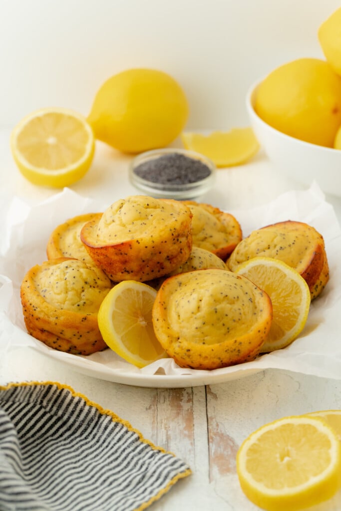 A plate of Bakery-Style Lemon Poppy Seed Muffins garnished with lemon slices, surrounded by whole and halved lemons, a bowl of poppy seeds, and a striped cloth on a light wooden surface.