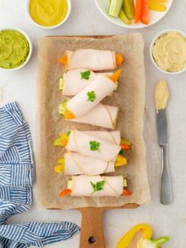 Four turkey and veggie roll-ups garnished with parsley are arranged on parchment paper atop a wooden board. Surrounding them are bowls of hummus, guacamole, mustard, and sliced vegetables. A blue-striped cloth is nearby.