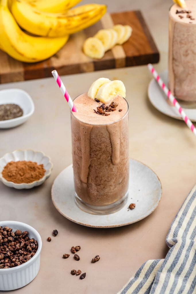A tall glass of healthy, high-protein chocolate smoothie topped with banana slices and cacao nibs, with a pink-striped straw. Ingredients like bananas, chia seeds, and cocoa powder are arranged around on a beige surface.