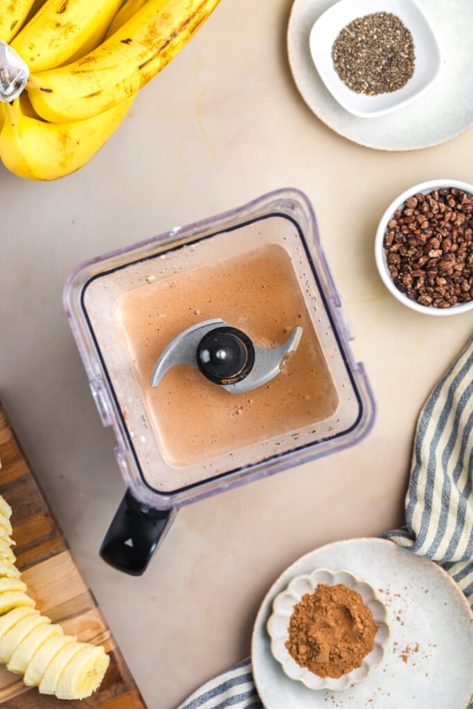 A blender with a creamy brown Healthy High-Protein Chocolate Smoothie sits on a countertop surrounded by bananas, chia seeds, cacao nibs, sliced bananas, and cocoa powder. A striped cloth is visible on the side.