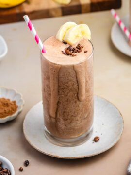 A tall glass of healthy, high-protein chocolate smoothie topped with banana slices and cacao nibs, with a pink-striped straw. Ingredients like bananas, chia seeds, and cocoa powder are arranged around on a beige surface.