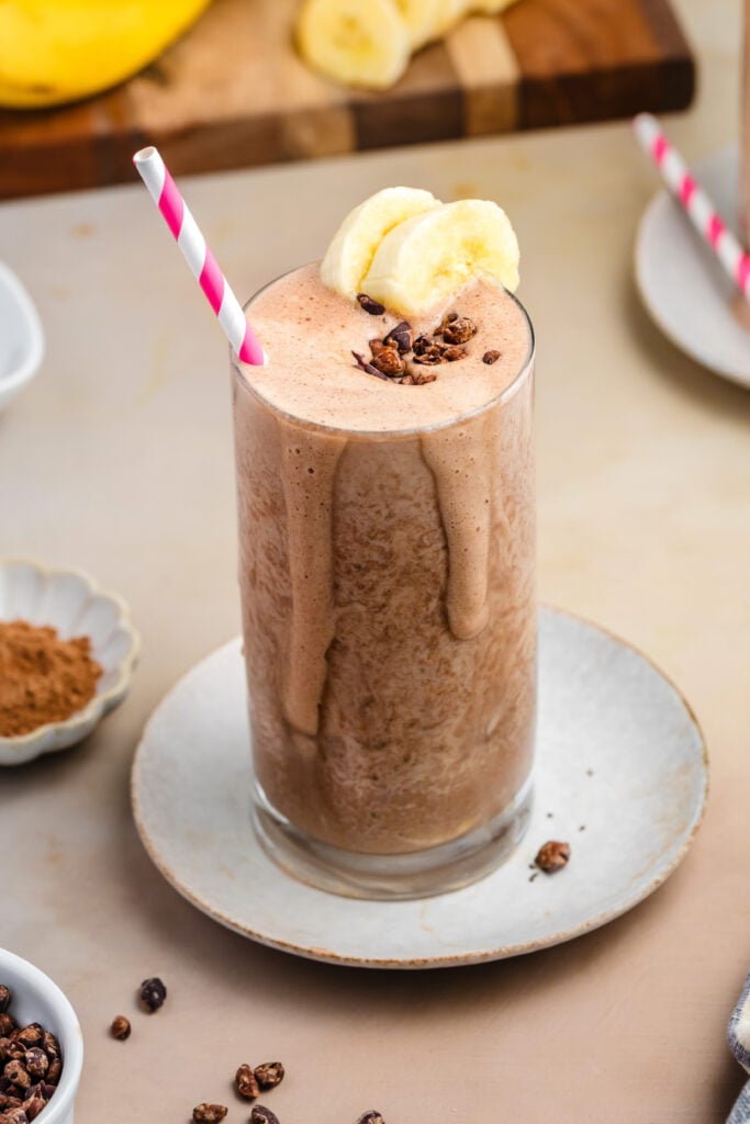 A tall glass of healthy, high-protein chocolate smoothie topped with banana slices and cacao nibs, with a pink-striped straw. Ingredients like bananas, chia seeds, and cocoa powder are arranged around on a beige surface.