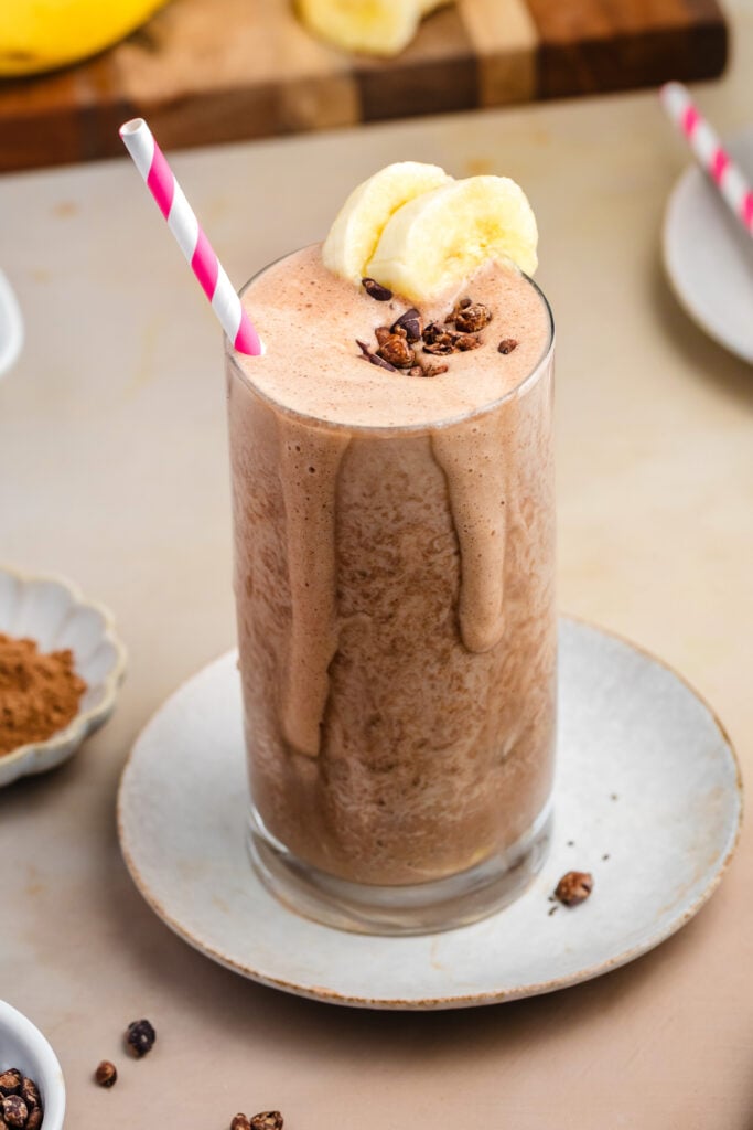 A tall glass of healthy, high-protein chocolate smoothie topped with banana slices and cacao nibs, with a pink-striped straw. Ingredients like bananas, chia seeds, and cocoa powder are arranged around on a beige surface.