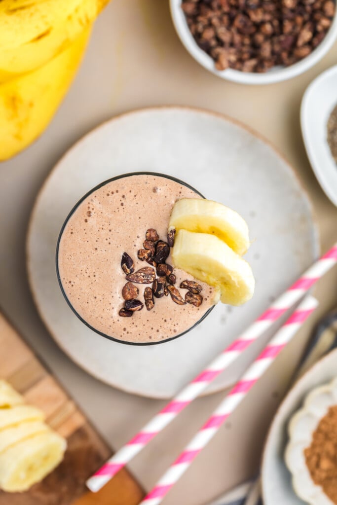 A healthy, high-protein chocolate smoothie topped with cacao nibs and banana slices, garnished with two pink-striped straws. No added sugar—just wholesome ingredients and delicious flavor in every sip. Banana slices are visible in the background.