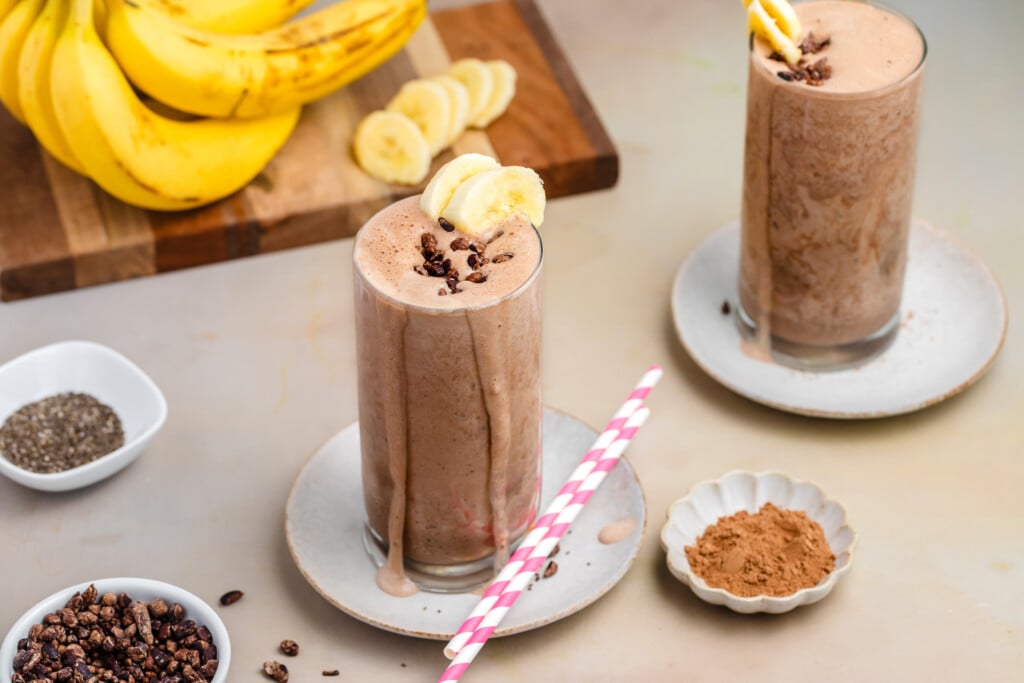 Two tall glasses of healthy chocolate banana smoothie, with no added sugar and high-protein goodness, are topped with banana slices and cacao nibs. Bananas, chia seeds, cacao nibs, and cocoa powder surround the drinks; a pink-striped straw lies nearby.