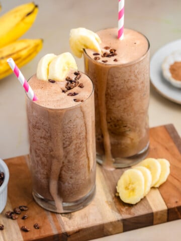 Two tall glasses of healthy chocolate banana smoothies with no added sugar and cacao nibs sit on a wooden board. Surrounded by bananas, cocoa powder, and more cacao nibs—it's the perfect high-protein treat.