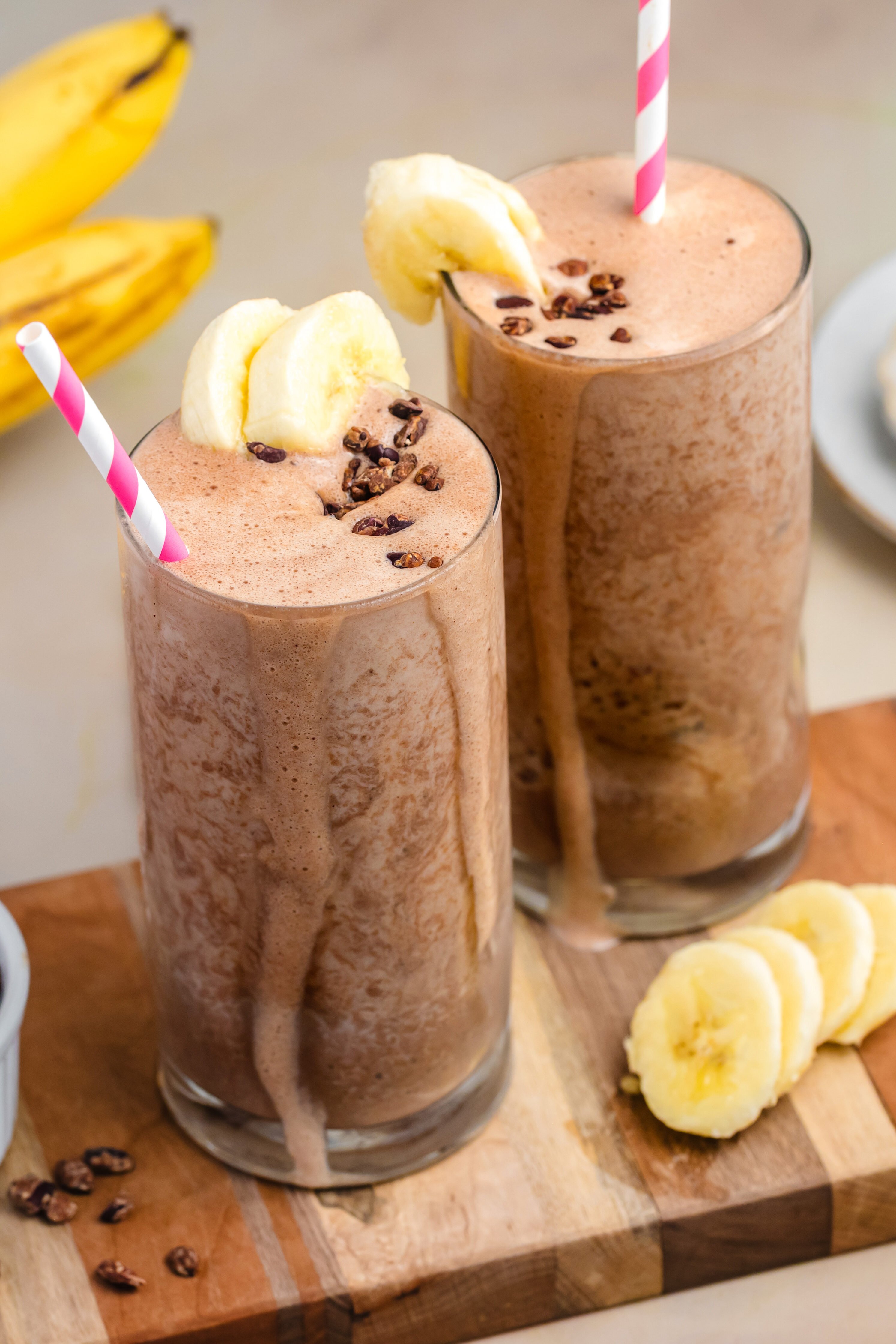 Two tall glasses of healthy chocolate banana smoothies with no added sugar and cacao nibs sit on a wooden board. Surrounded by bananas, cocoa powder, and more cacao nibs—it's the perfect high-protein treat.