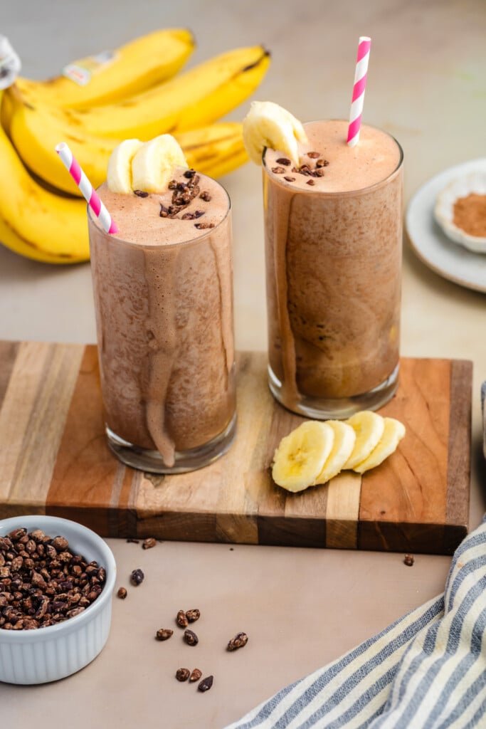 Two glasses of healthy, high-protein chocolate banana smoothies with pink-striped straws, garnished with banana slices and cocoa nibs, are placed on a wooden board. Bananas and a small bowl of cocoa nibs are nearby.