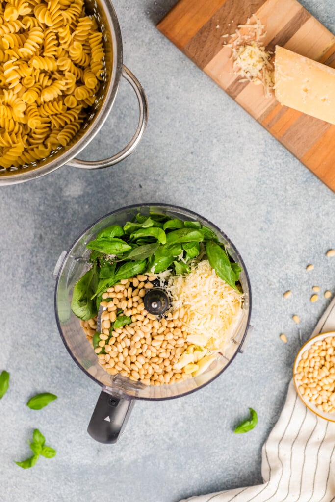 A food processor filled with basil, pine nuts, shredded cheese, and garlic sits on a counter next to a pot of cooked pasta for Pesto Caprese Pasta Salad. Pine nuts and basil leaves are scattered nearby alongside a cutting board with a wedge of cheese.