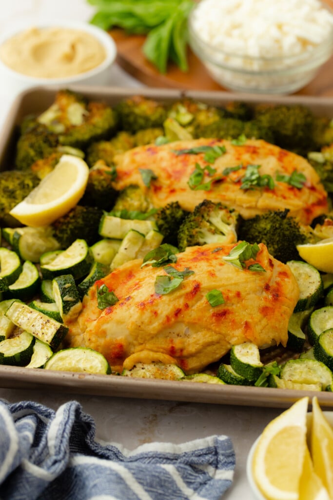 Roasted Garlic Hummus Chicken & Veggies (Easy One Sheet Dinner): Baked chicken breasts topped with herbs, surrounded by roasted broccoli and zucchini, and garnished with lemon wedges. A blue-striped cloth and bowls of dip, basil, and cheese complete the scene.
