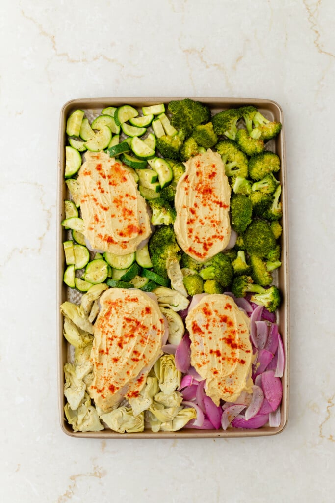 A sheet pan with three pieces of Roasted Garlic Hummus Chicken & Veggies (Easy One Sheet Dinner), topped with hummus and paprika, surrounded by chopped zucchini, broccoli, red onion, and artichoke hearts on a light-colored surface.