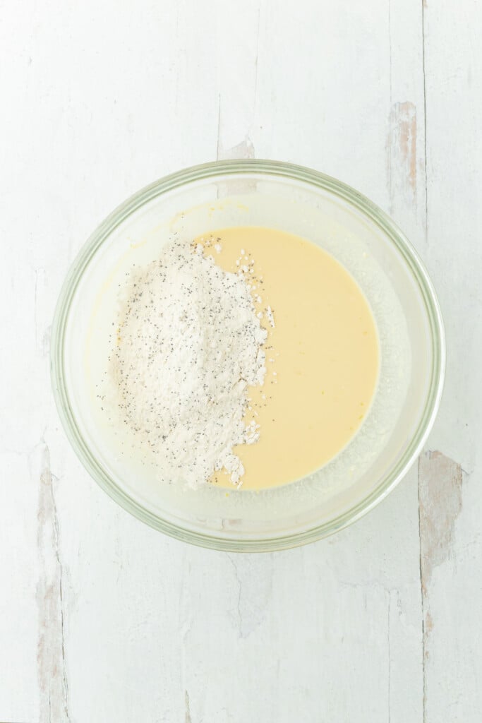 A glass bowl with a yellowish batter and a mound of flour topped with black pepper, set on a white wooden surface—perfect for making bakery-style lemon poppy seed muffins.
