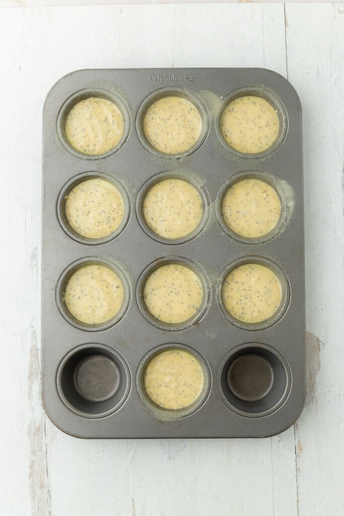 A 12-cup muffin pan filled with pale yellow Bakery-Style Lemon Poppy Seed Muffins batter, speckled with poppy seeds and two cups empty, sits on a light-colored surface.