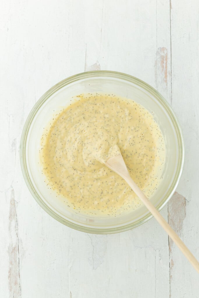 A glass bowl filled with Bakery-Style Lemon Poppy Seed Muffins batter sits on a light wooden surface. A wooden spoon rests in the bowl, partially submerged in the creamy, speckled mixture.