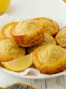 A white bowl filled with Bakery-Style Lemon Poppy Seed Muffins and lemon wedges, surrounded by whole and sliced lemons, a small bowl of poppy seeds, and a striped cloth on a white surface.