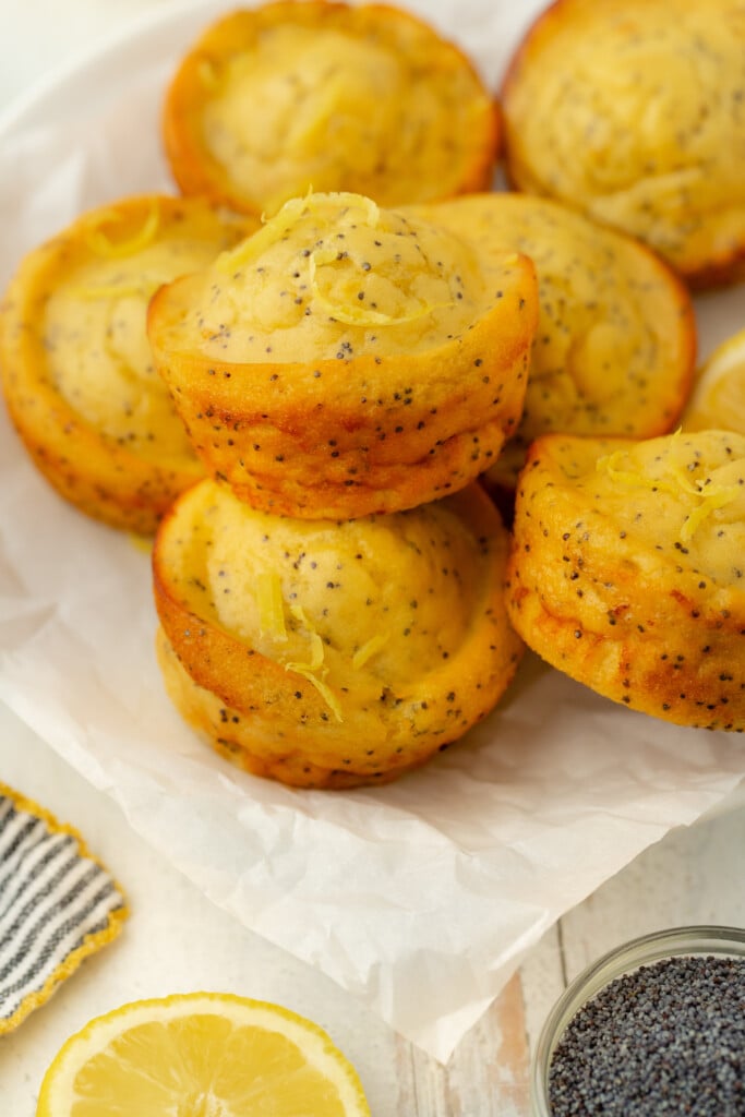 A stack of bakery-style lemon poppy seed muffins sits on white parchment paper, with visible poppy seeds and lemon zest. Slices of fresh lemon and a small bowl of poppy seeds are nearby.