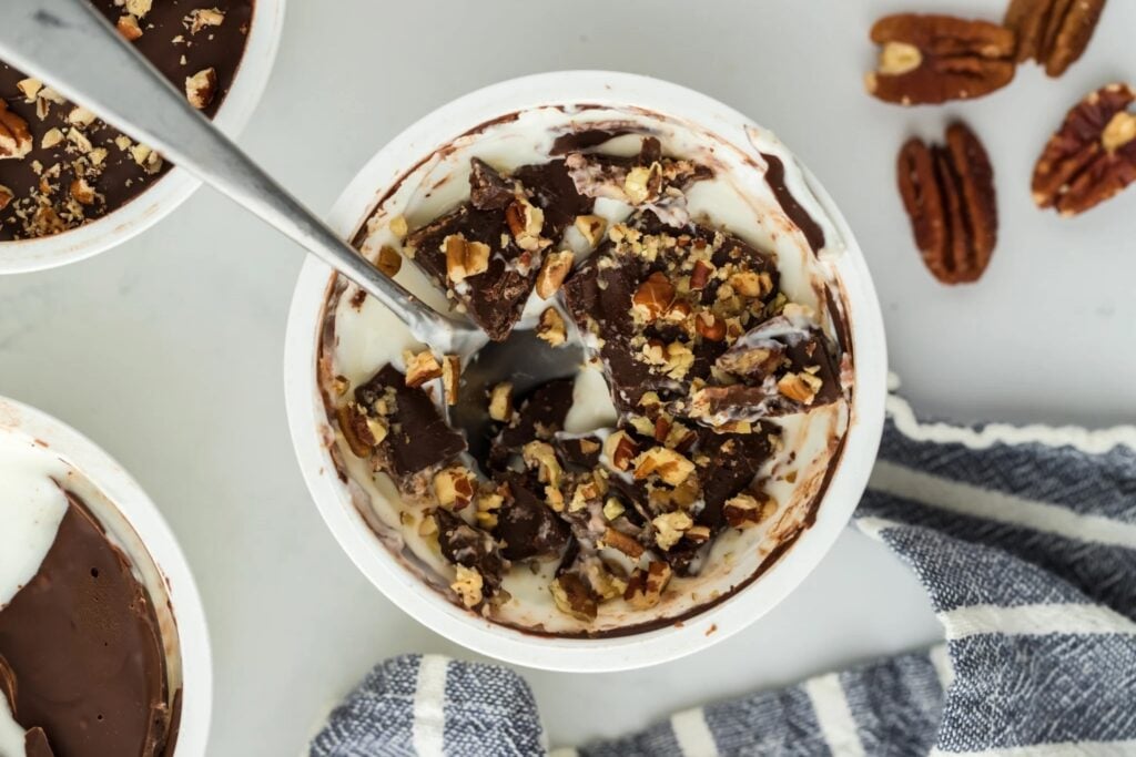 A bowl of creamy, high protein snack dessert topped with chopped pecans and chocolate, partially eaten with a spoon inside. A striped cloth and a few pecans are nearby on a light surface.